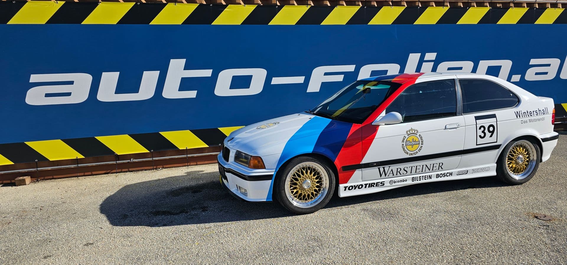 Weißer BMW 3er Compact in DTM-Renndekor mit blau-rotem M-Streifen, Startnummer 39 und goldenen Kreuzspeichenfelgen vor der blau-gelben Firmenplane von auto-folien.at