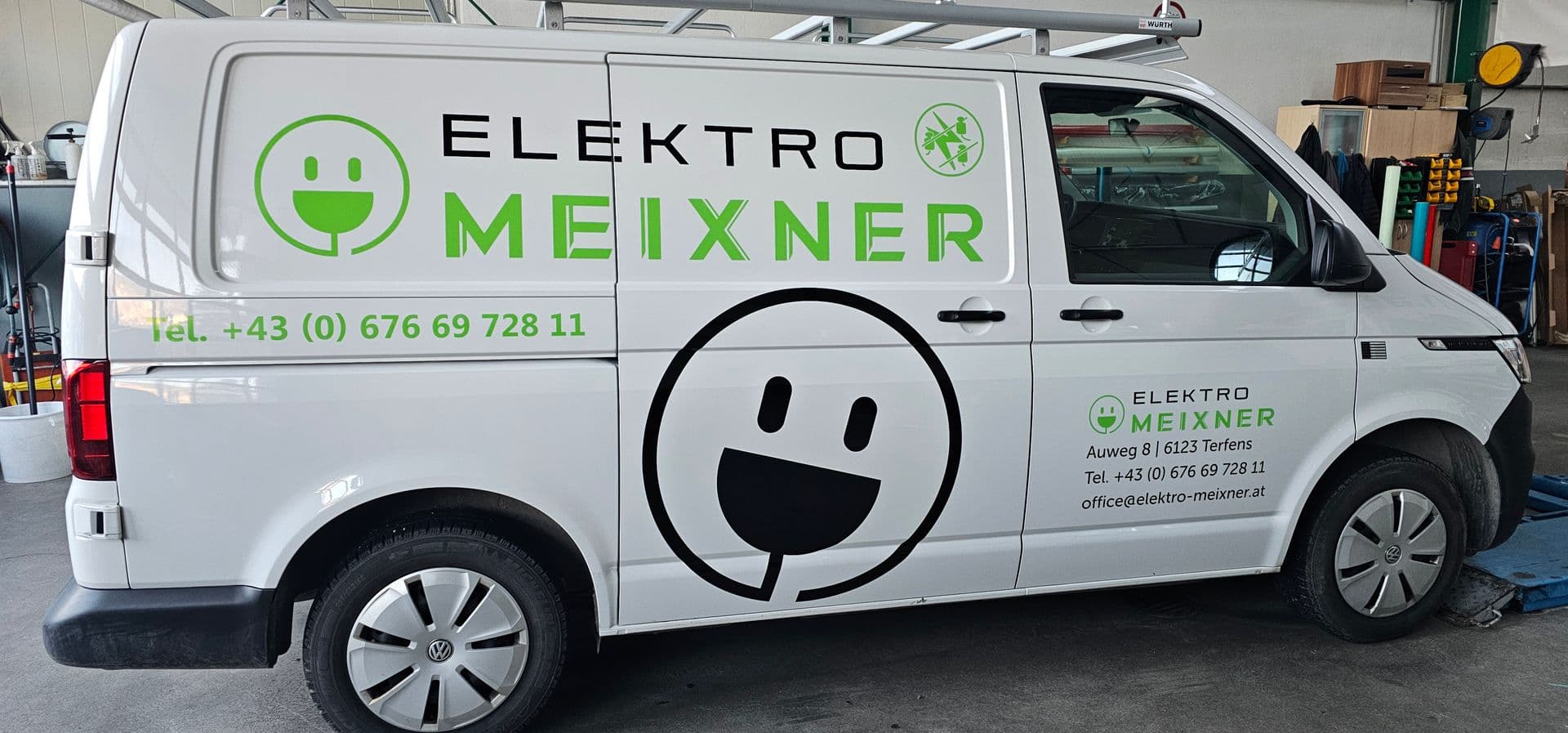 Weißer VW Transporter mit grünem Steckersymbol und Beschriftung eines Elektrobetriebs in der Werkstatt