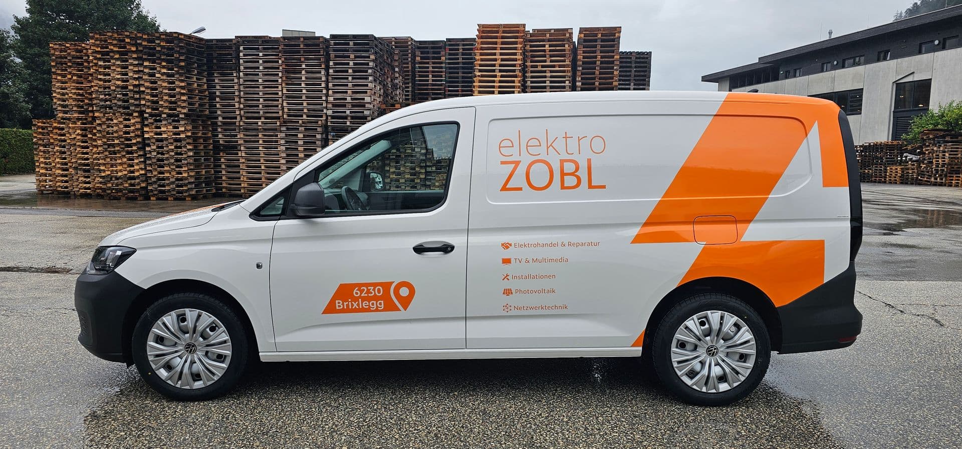 Weißer VW Caddy mit oranger Blitzgrafik und Leistungsübersicht eines Elektrobetriebs, dahinter Palettenstapel