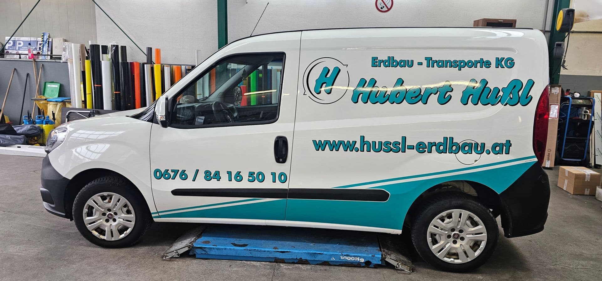 Weißer Fiat Doblò mit türkiser Beschriftung eines Erdbauunternehmens in der Werkstatt, links das Folienlager