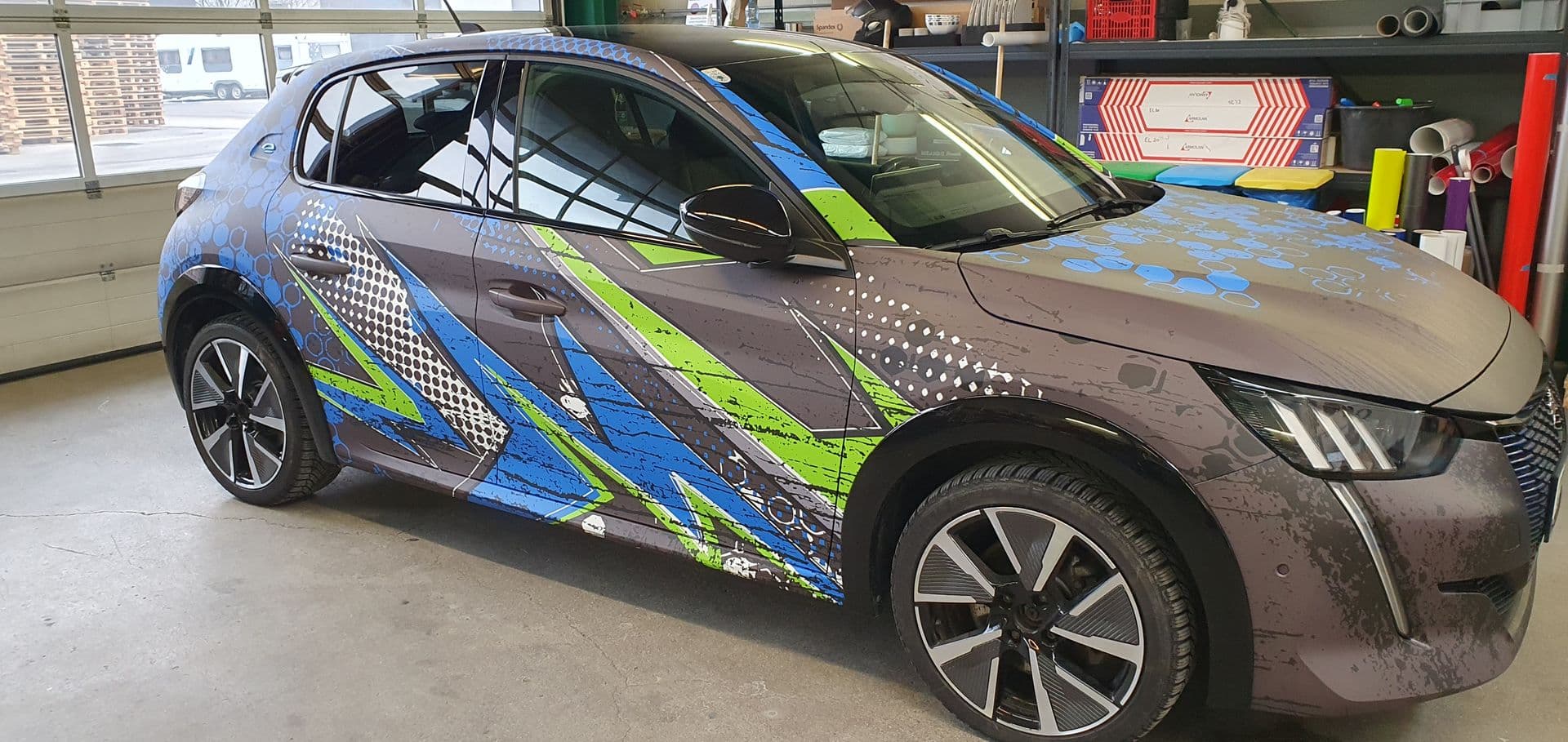 Peugeot e-208 mit blau-grüner Designfolierung über Flanke und Dach, in der Werkstatt