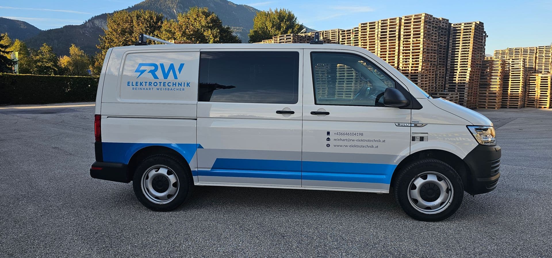 Weißer VW Transporter mit blauem Seitenstreifen und Logo eines Elektrotechnikbetriebs, dahinter Palettenstapel und Berge