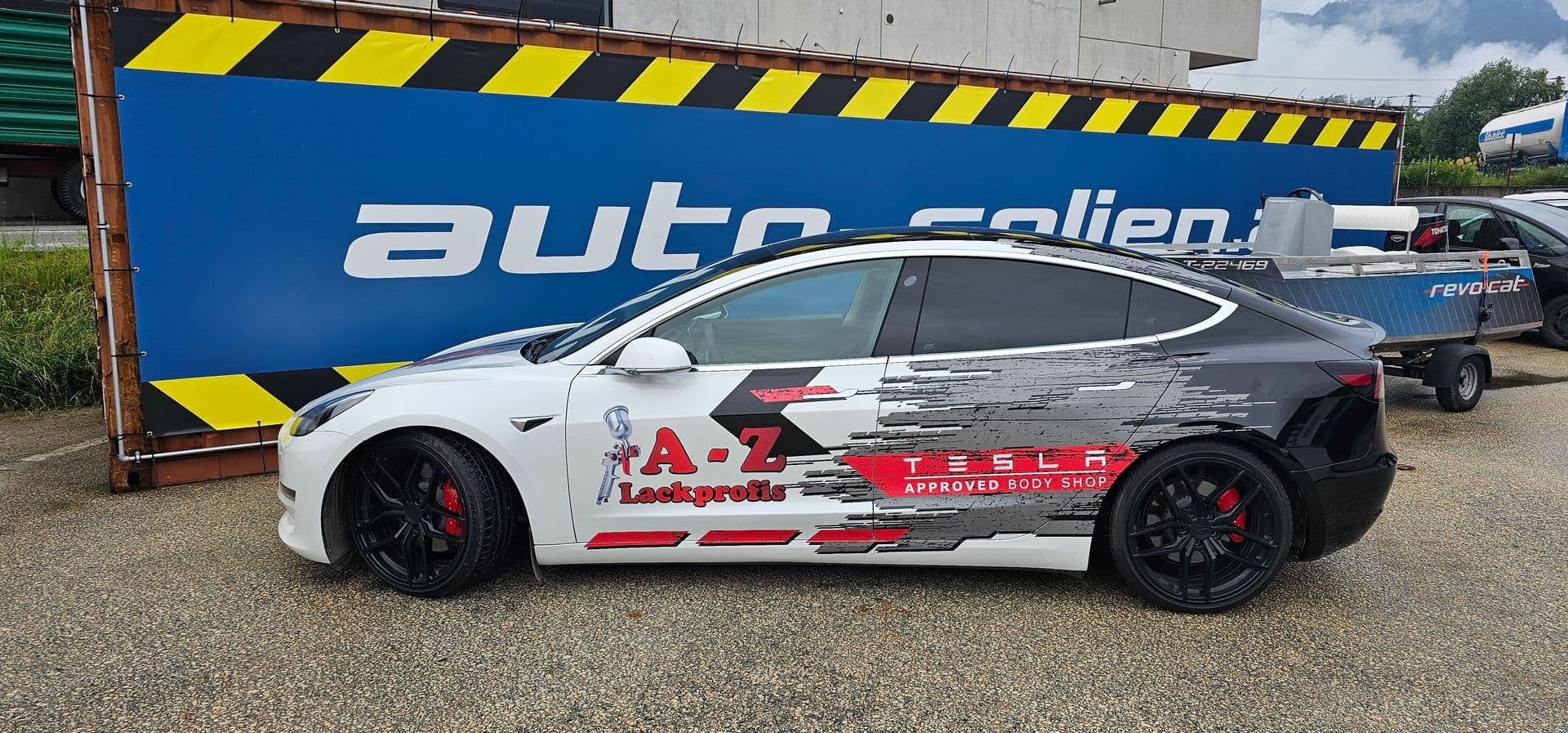 Weißer Tesla Model 3 mit grau-roter Splittergrafik und Beschriftung eines Lackierbetriebs vor der Firmenplane von auto-folien.at