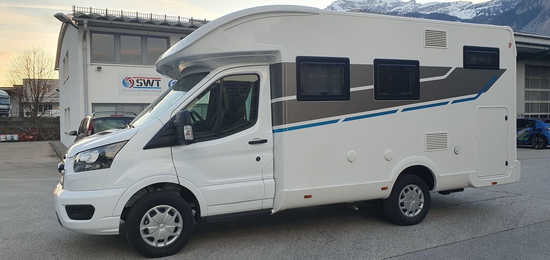 Weißes Wohnmobil mit grau-blauem Dekorstreifen vor bewaldeten Berghängen